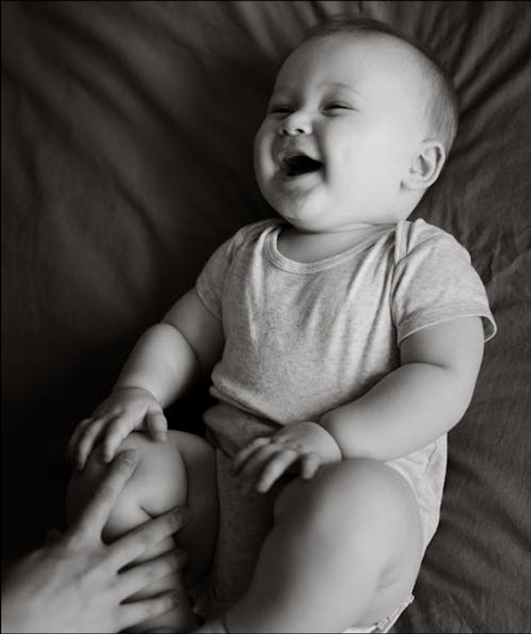 A black and white photo of a baby laughing at the touch of a hand on their knee.